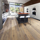 A modern kitchen interior with brown engineered wood flooring, an island with two white bar stools, white cabinetry and large windows.