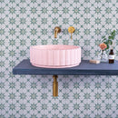 Pink sink on a blue shelf with gold fixtures against green patterned wall tiles