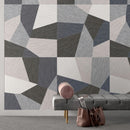 Geometric patterned wall tiles with textured fabric-like panels in gray, charcoal, off-white, and muted blue, with a grey bench, a pink cushion, and a brown leather bag.