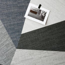 A close-up of a geometric fabric effect tile with textured woven sections in charcoal gray, light gray, and slate blue. A small printed postcard is on top of the tile.