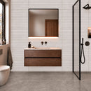 Bathroom with a wall mounted dark wood vanity, a large black-framed square mirror,  light grey stone effect wall tiles and darker grey floor tiles.