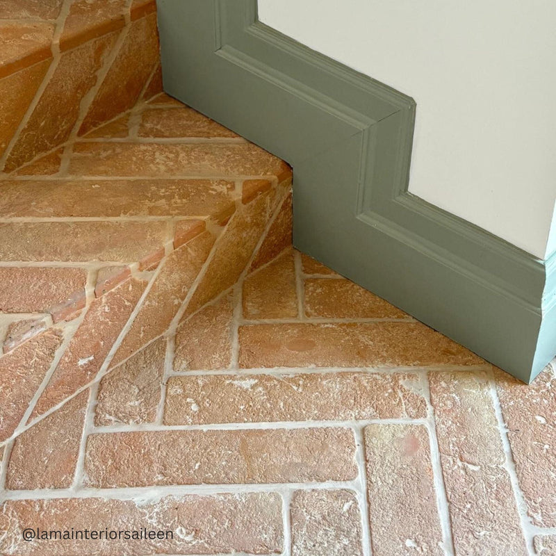 Close-up of a terracotta stone floor with a green skirting board and white wall.