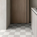 A kitchen with a checkered gre y and white tile floor, a tall brown wooden cupboard and a pale worktop.