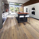 A modern kitchen interior with brown engineered wood flooring, an island with two white bar stools, white cabinetry and large windows.