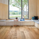 Light natural wood plank flooring in a bright room with a cushioned wooden bench by a large window.
